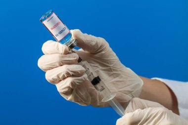 Tıbbi eldiven giyen bir doktor COVID-19 aşısı olan bir test tüpünden ilaç alıyor ve mavi arka planda bir şırıngaya 