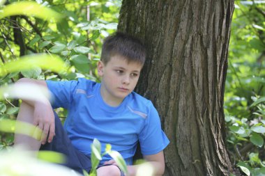 Bir gencin parkta yürürken bir fotoğrafçı için poz veriyor. Oturur, ağaca yaslanmış.