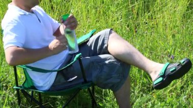 Bir adam bir ışıklı çayır piknik sandalyeye oturur. Duruyor, bir akıllı telefon üzerinde çalışıyor, su içiyor. Bitkiler ve çiçekler yaz rüzgardan sway.