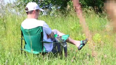 Bir adam bir ışıklı çayır piknik sandalyeye oturur. O duruyor, sonra bırakır. Bitkiler ve çiçekler yaz rüzgardan sway.