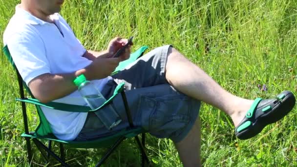 明るい草原のピクニック椅子に座っています 彼にかかっている スマート フォン上で動作し 葉します ハーブや花が夏の風から動揺します — ストック動画