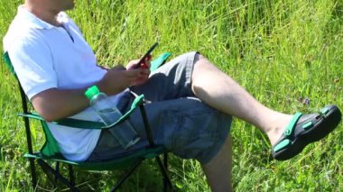 Bir adam bir ışıklı çayır piknik sandalyeye oturur. O istirahat, bir akıllı telefon üzerinde çalışıyor. Bitkiler ve çiçekler yaz rüzgardan sway.