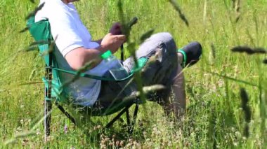 Bir adam bir katlanır piknik sandalyede oturur ve bir smartphone ile çalışır. O zaman kadar alır ve bırakır. Çevresinde ot, Rüzgar tarafından eğik el sallıyor