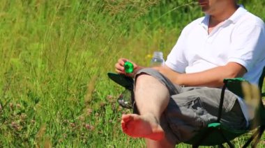 Bir adam bir katlanır piknik sandalyede oturur ve su içiyor. Şişe kapatır ve stand koyar.