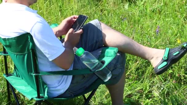 一个男人坐在一个被点燃的草地上的野餐椅上 他在休息 在智能手机上工作 香草和花朵从夏日的风中摇摆 — 图库视频影像