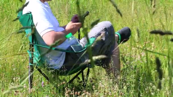 男は折り畳みピクニック椅子に座っている スマート フォンで動作します その後 彼は起き 周りが振っている草 風に傾いています — ストック動画