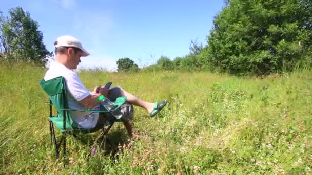 男は折り畳みピクニック椅子に座っている スマート フォンで動作します 周りが振っている草 風に傾いています — ストック動画