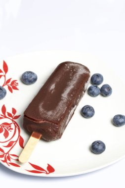 Dondurma buz çikolata bir sopa. Kırmızı süslemeli beyaz bir plaka üzerinde yatıyor. Yaban mersini yanlarında vardır. Beyaz arka plan.