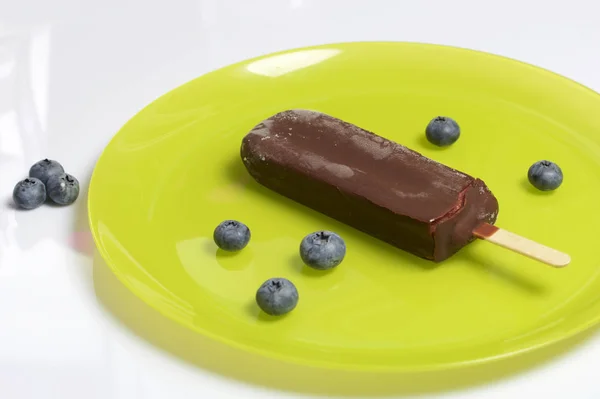 Dondurma buz çikolata bir sopa. Açık yeşil renkli bir tabağa yatıyor. Yaban mersini yanlarında vardır. Beyaz arka plan.