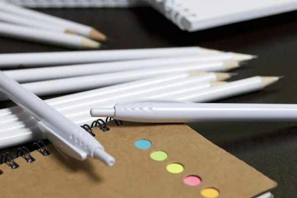 White spring pads, simple pencils and ballpoint pens for notes and sketches. Near a notebook of a different color. Stationery for school and teaching. On a dark background.