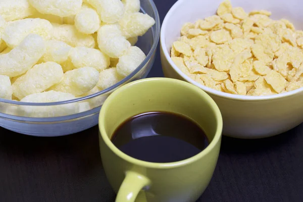 Useful breakfast. Corn sticks flakes in a container and a mug of coffee. On a dark background.