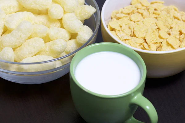Useful breakfast. Corn sticks flakes in a container and a mug of milk. On a dark background.