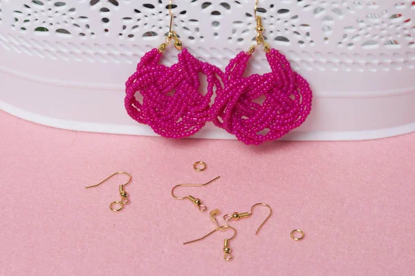 Earrings from beads handmade. Pink colour. Hang on the stand. Nearby are scattered ear wires and other accessories. Needlework at home. Bead jewelery.