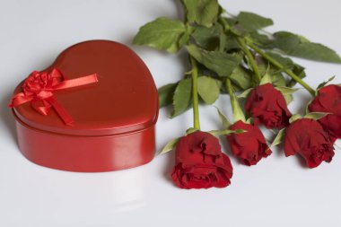 Bir buket kırmızı gül ve kalp şeklinde bir kutu içinde bir hediye. Beş çiçek beyaz bir yüzeye yalan.