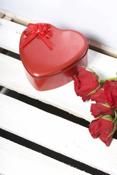 A bouquet of scarlet roses and a gift in a box in the shape of a heart. Five flowers lie on a wooden box. On a white background.