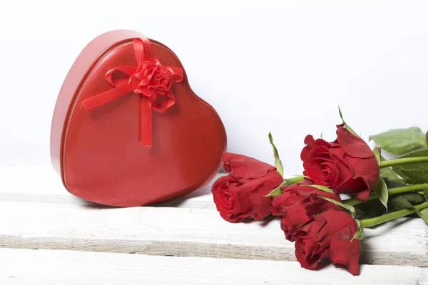 A bouquet of scarlet roses and a gift in a box in the shape of a heart. Five flowers lie on a wooden box. On a white background.