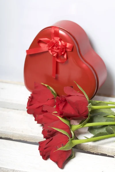 A bouquet of scarlet roses and a gift in a box in the shape of a heart. Five flowers lie on a wooden box. On a white background.