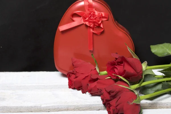 A bouquet of scarlet roses and a gift in a box in the shape of a heart. Flowers lie on a wooden box. On a black background.