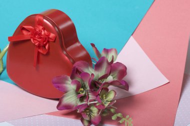 Bir hediye ile kırmızı bir kalp şeklinde bir kutu. Sonraki bir çiçektir. Çok renkli kağıt bir arka plan üzerinde.