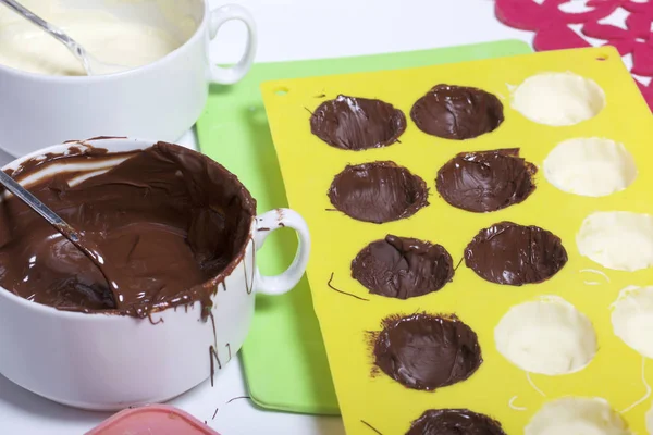 Melted white and black chocolate in white cups. Nearby silicone mold smeared with chocolate.