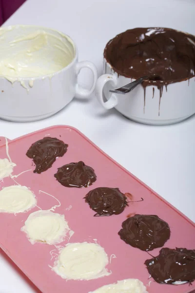 Melted white and black chocolate in white cups. Nearby silicone mold smeared with chocolate.