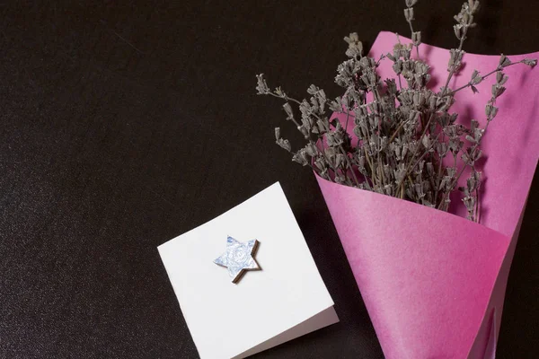 A bouquet of dried flowers, wrapped in colored paper. Near a small greeting card. On a dark background.
