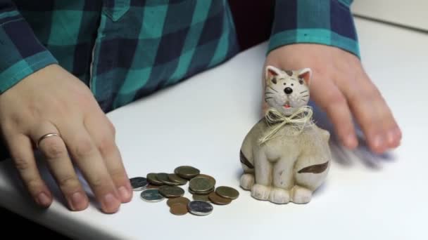 テーブルの上は猫の形の貯金箱銀行です 男がテーブルからコインをピックアップし 貯金箱に入れます — ストック動画