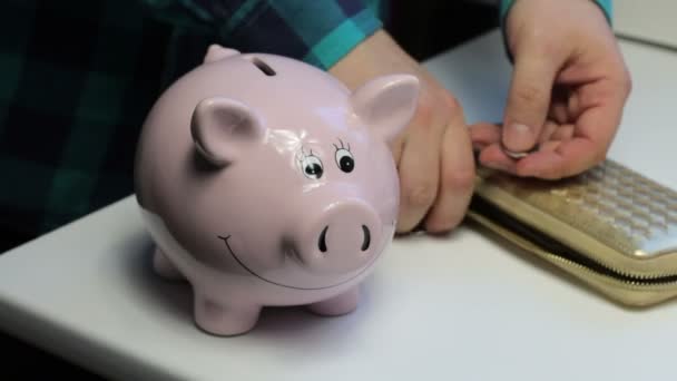 桌子上有一个粉红色猪形状的存钱罐 说谎的钱包和散落的硬币 一个男人从桌子上收集硬币 放在存钱罐里 然后关上钱包 — 图库视频影像