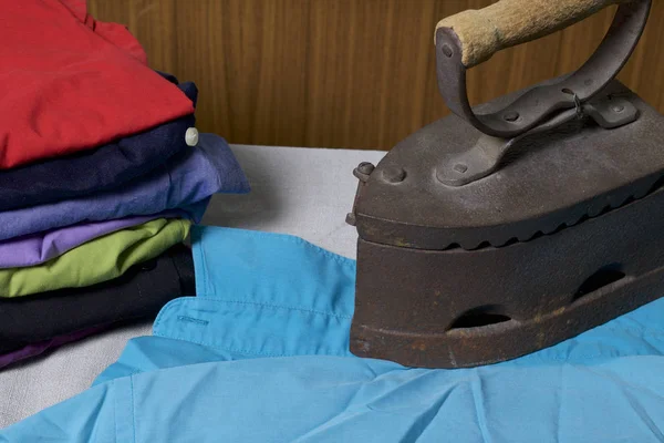 Old iron, heated by hot coals. Located on linen fabric. Standing on an ironing board. Beside a stack of undressed shirts.