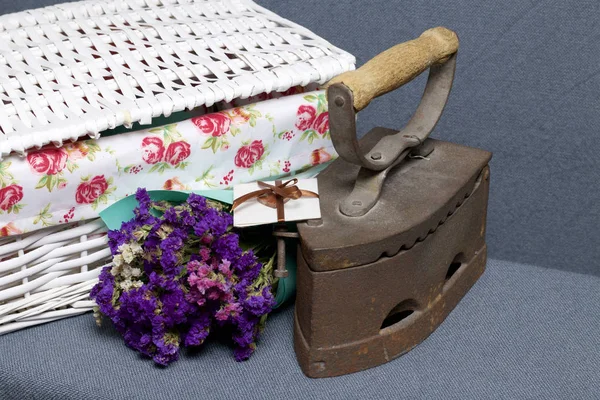 Old iron, heated by hot coals. Located on gray fabric. Nearby are wicker baskets, a bouquet of dried flowers and a greeting card.