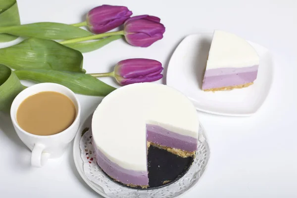 Blueberry cheesecake. A piece of ready-made dessert on a saucer. Jelly layers of different colors are visible. Near a cup of coffee and a bouquet of tulips. — Stock Photo, Image