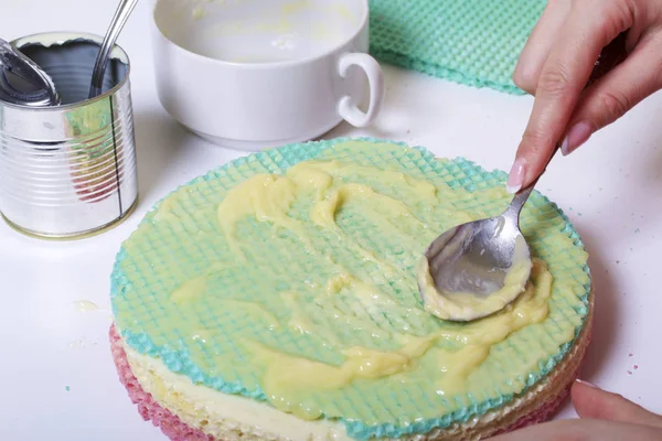 Una mujer lubrica los pasteles y los pone en una pila. Tortas redondas de obleas de diferentes colores. Por hacer tarta de gofres. Cerca hay una lata de leche condensada para remojar los pasteles . — Foto de Stock
