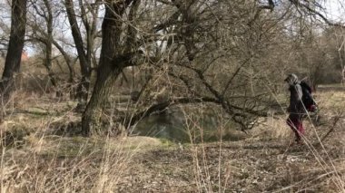 Bir erkekte bir kaputa ceketle nehir kıyısında yürüyor. Bir plastik şişe yükseltir, onunla çöp alır. Bahar manzara. Çevre koruma.