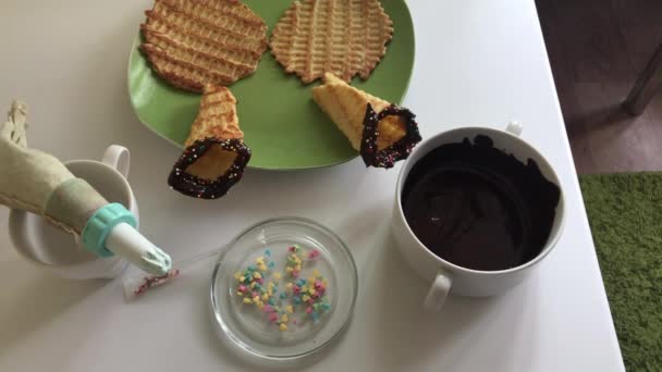 桌子上躺着一个华夫饼锥 浸在融化的巧克力和装饰与多色的洒水 有烹饪用的食材 — 图库视频影像