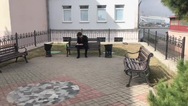 若い男性がベンチに座り ノートにスケッチを作ります 涼しい天気 男は黒いコートを着ています レトロなスタイルの居心地の良い中庭 — ストック動画