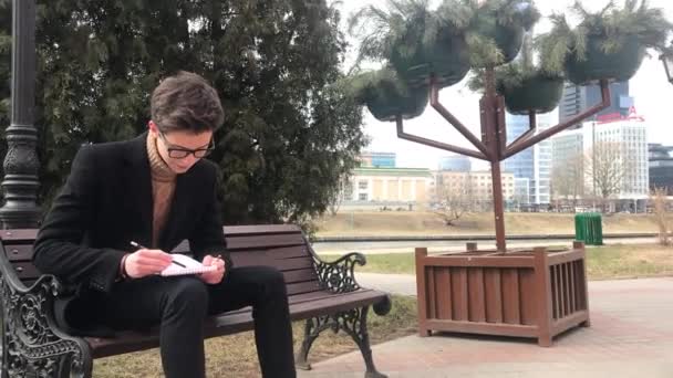 若い男性がベンチに座り ノートにスケッチを作ります ページを回転します 涼しい天気 男は黒いコートを着ている — ストック動画