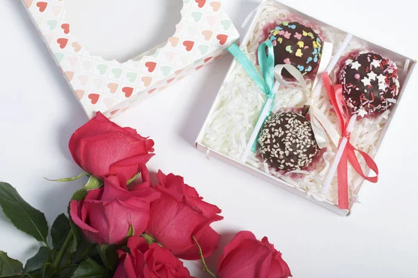 Cake pops are beautifully packed in a gift box. Nearby there is a cover with a transparent window and a bouquet of scarlet roses.
