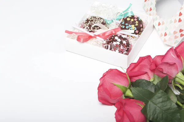 Cake pops are beautifully packed in a gift box. Nearby there is a cover with a transparent window and a bouquet of scarlet roses.