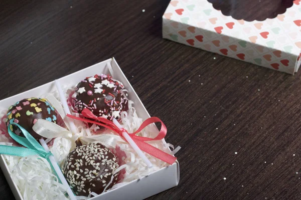Cake pops are beautifully packed in a gift box. Nearby there is a cover with a transparent window. On a dark background.