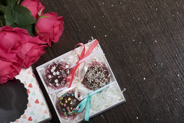 Cake pops are beautifully packed in a gift box. Nearby there is a cover with a transparent window and a bouquet of scarlet roses. On a dark background.