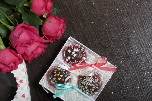Cake pops are beautifully packed in a gift box. Nearby there is a cover with a transparent window and a bouquet of scarlet roses. On a dark background.