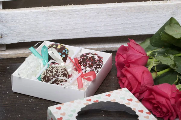 Los pastelitos están bellamente empaquetados en una caja de regalo. Cerca hay una cubierta con una ventana transparente y un ramo de rosas escarlata. Sobre el fondo de tablas de madera — Foto de Stock