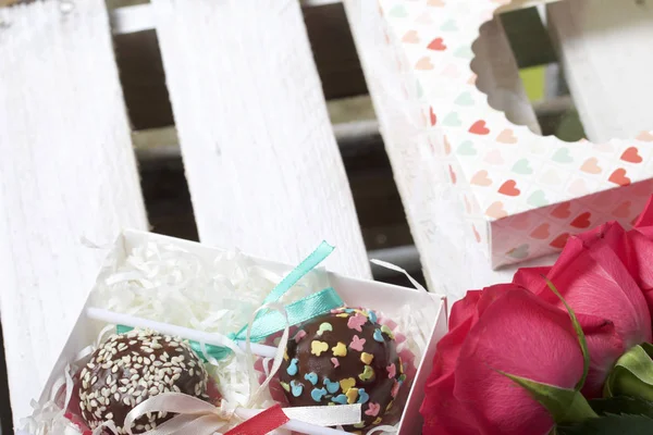 Cake pops are beautifully packed in a gift box. Nearby there is a cover with a transparent window and a bouquet of scarlet roses. On the background of wooden boards
