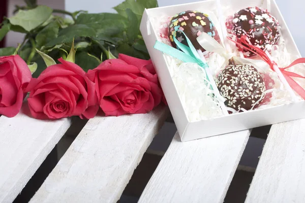 Cake pops are beautifully packed in a gift box. Nearby is a bouquet of scarlet roses. On the background of wooden boards