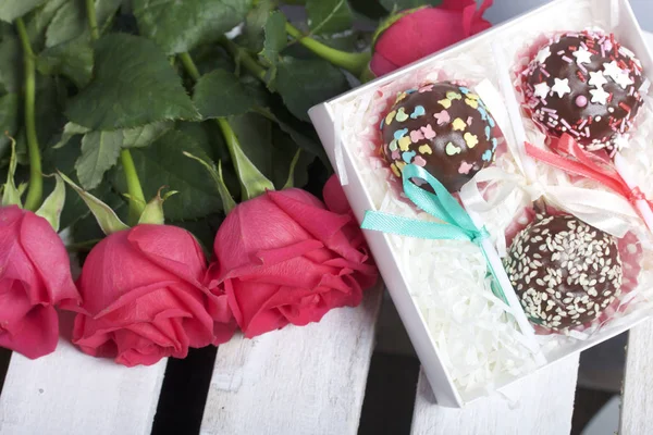 Cake pops are beautifully packed in a gift box. Nearby is a bouquet of scarlet roses. On the background of wooden boards