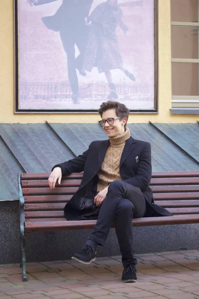 Un jeune homme avec un sourire assis sur un banc. Habillé d'un manteau léger noir, lunettes à bord noir . — Photo