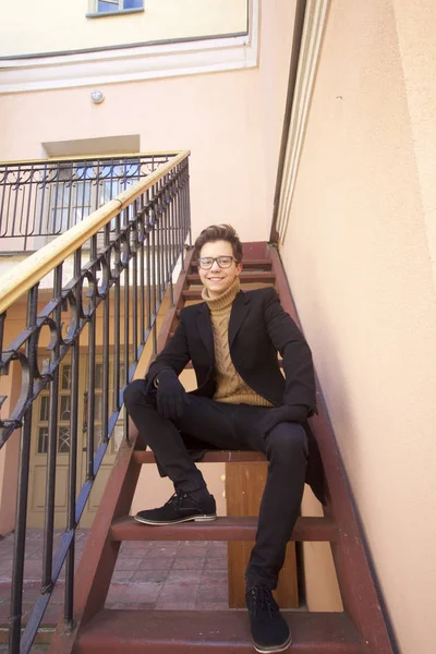 Un jeune homme au sourire pose assis sur un escalier vintage dans un style rétro. Habillé d'un manteau léger noir, lunettes à bord noir . — Photo