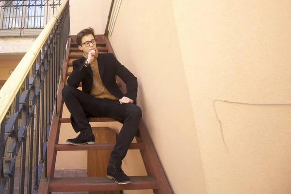 Un jeune homme au sourire pose assis sur un escalier vintage dans un style rétro. Habillé d'un manteau léger noir, lunettes à bord noir . — Photo