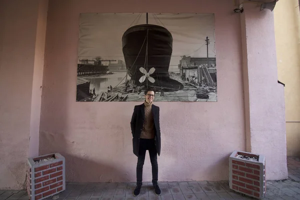 Un jeune homme avec un sourire pose près du mur de la ville, avec une image de style rétro. Habillé d'un manteau léger noir, lunettes à bord noir . — Photo