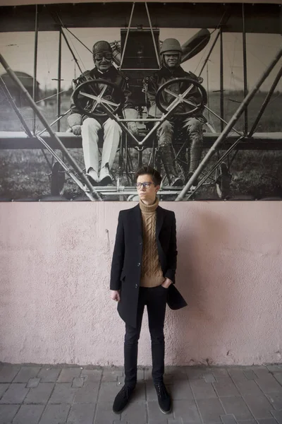 Un jeune homme avec un sourire pose près du mur de la ville, avec une image de style rétro. Habillé d'un manteau léger noir, lunettes à bord noir . — Photo